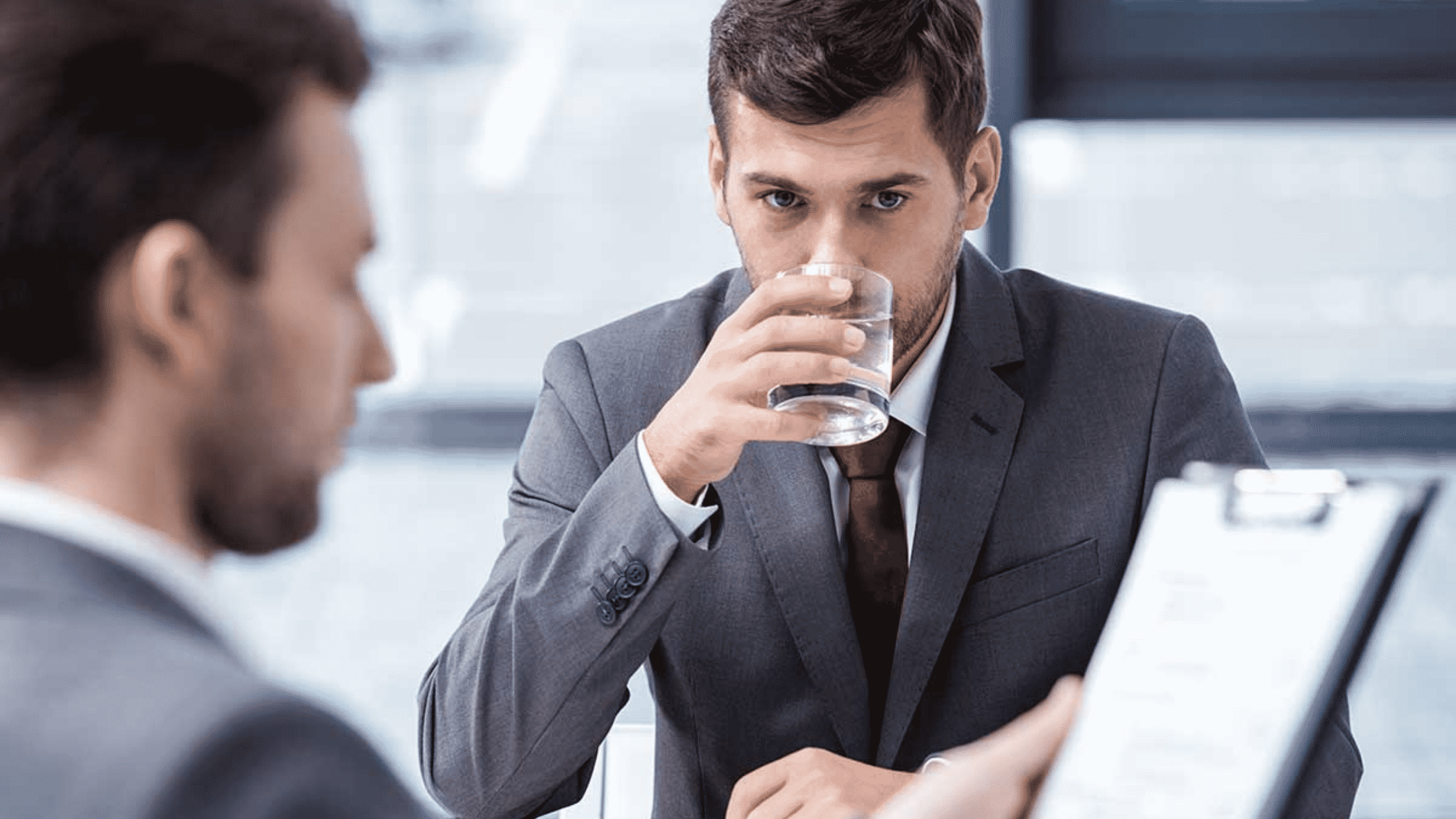 Homme buvant un verre d'eau en face de son recruteur qui regarde son CV durant un entretien d'embauche