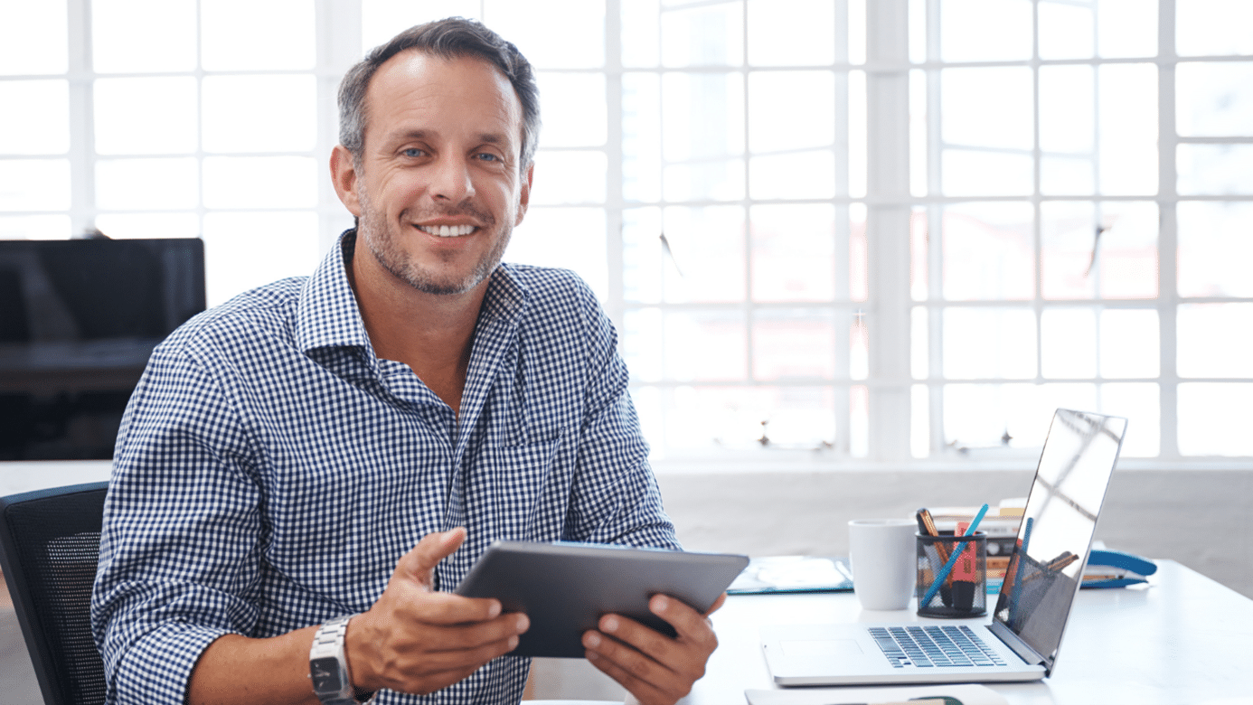 Un homme qui sourit en tenant une tablette dans la main, a côté de son bureau pour illustrer le top 6 des tendances de recrutement à surveiller pour la rentrée